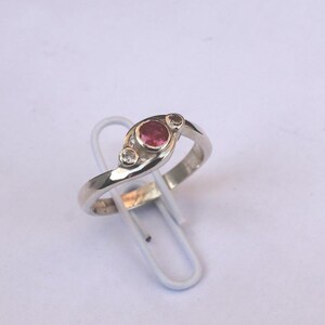 Könnte beinhalten: Ein silberfarbener Ring mit einem zentralen runden rosa Edelstein, der von zwei kleineren klaren Steinen flankiert wird. Die Ringschiene hat eine glatte, polierte Oberfläche und ein geschwungenes Design, das die drei Steine in einer Zargenfassung hält.