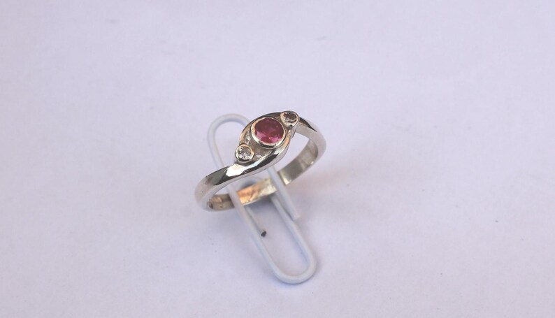 K&ouml;nnte beinhalten: Ein silberfarbener Ring mit einem zentralen runden rosa Edelstein, der von zwei kleineren klaren Steinen flankiert wird. Die Ringschiene hat eine glatte, polierte Oberfl&auml;che und ein geschwungenes Design, das die drei Steine in einer Zargenfassung h&auml;lt.