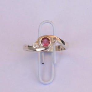 K&ouml;nnte beinhalten: Ein silberfarbener Ring mit einem zentralen runden rosa Edelstein, der von zwei kleineren klaren runden Steinen flankiert wird. Die Ringschiene hat eine glatte, polierte Oberfl&auml;che und ein &uuml;berlappendes Design, das die drei Steine h&auml;lt.