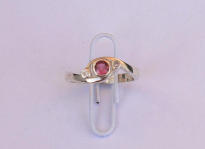 K&ouml;nnte beinhalten: Ein silberfarbener Ring mit einem zentralen runden rosa Edelstein, der von zwei kleineren klaren runden Steinen flankiert wird. Die Ringschiene hat eine glatte, polierte Oberfl&auml;che und ein &uuml;berlappendes Design, das die drei Steine h&auml;lt.