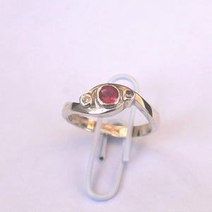 K&ouml;nnte beinhalten: Ein silberfarbener Ring mit einem zentralen, oval geschliffenen rosa Edelstein, der von zwei kleineren, klaren, rund geschliffenen Steinen flankiert wird. Die Ringschiene ist glatt poliert und in einem geschwungenen Design gehalten.