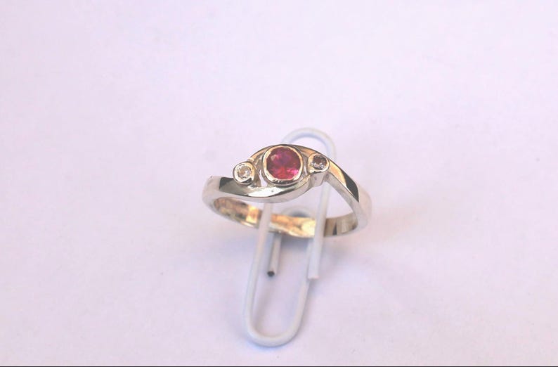 K&ouml;nnte beinhalten: Ein silberfarbener Ring mit einem zentralen, oval geschliffenen rosa Edelstein, der von zwei kleineren, klaren, rund geschliffenen Steinen flankiert wird. Die Ringschiene ist glatt poliert und in einem geschwungenen Design gehalten.