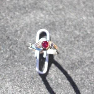 K&ouml;nnte beinhalten: Ein silberfarbener Ring mit einem zentralen, rund geschliffenen roten Edelstein, der von zwei kleineren, klaren, rund geschliffenen Steinen flankiert wird. Die Ringschiene hat ein Bypass-Design, bei dem sich die Enden &uuml;berlappen.
