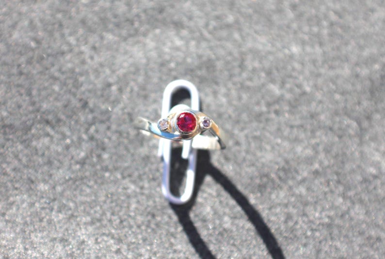 K&ouml;nnte beinhalten: Ein silberfarbener Ring mit einem zentralen, rund geschliffenen roten Edelstein, der von zwei kleineren, klaren, rund geschliffenen Steinen flankiert wird. Die Ringschiene hat ein Bypass-Design, bei dem sich die Enden &uuml;berlappen.