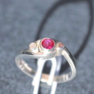 K&ouml;nnte beinhalten: Ein silberfarbener Ring mit einem zentralen, kreisf&ouml;rmigen, leuchtend rosa Edelstein in einer Fassung, flankiert von zwei kleineren, helleren runden Steinen auf beiden Seiten. Die Ringschiene hat eine glatte, polierte Oberfl&auml;che und ein asymmetrisches Design.