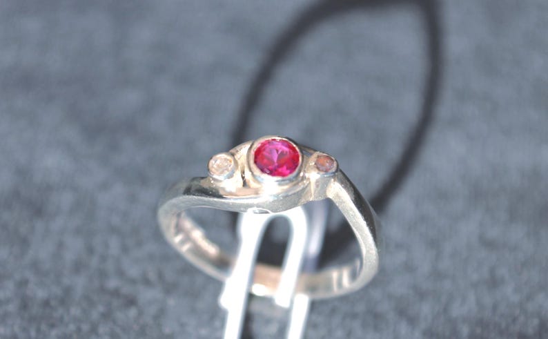 K&ouml;nnte beinhalten: Ein silberfarbener Ring mit einem zentralen, kreisf&ouml;rmigen, leuchtend rosa Edelstein in einer Fassung, flankiert von zwei kleineren, helleren runden Steinen auf beiden Seiten. Die Ringschiene hat eine glatte, polierte Oberfl&auml;che und ein asymmetrisches Design.