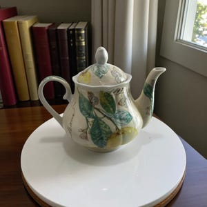 以下が含まれることがあります： クリーム色のセラミックティーポット。蓋、取っ手、注ぎ口付き。緑の葉、黄色のレモン、蝶が描かれています。ティーポットは白い表面に置かれています。