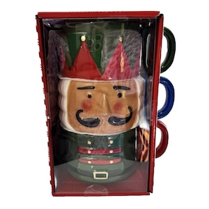 May include: A set of three ceramic mugs in a red box. The mugs are stacked and feature a nutcracker design with a red crown, a tan face, and a black mustache. The handles are green, blue, and red.