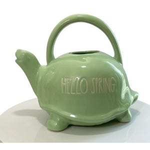 Puede incluir: Una regadera de cerámica verde claro con forma de tortuga y un asa curva. Las palabras "HELLO SPRING" están grabadas en relieve en el lateral. La regadera tiene un pico y se apoya en tres patas pequeñas.