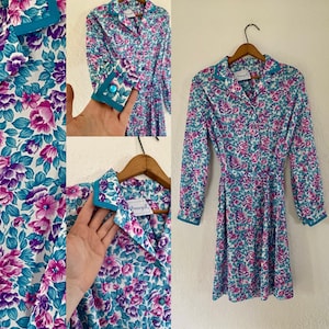 May include: A vintage floral dress with a button-down front, long sleeves, and a matching belt. The dress features a vibrant pattern of pink and purple flowers with teal accents on a white background. The collar and cuffs are trimmed with teal.