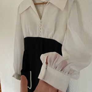 May include: A white and black dress with a large collar and long sleeves. The dress has a white top with pearl buttons and a black skirt. The sleeves are sheer and have a ruffle at the cuff.