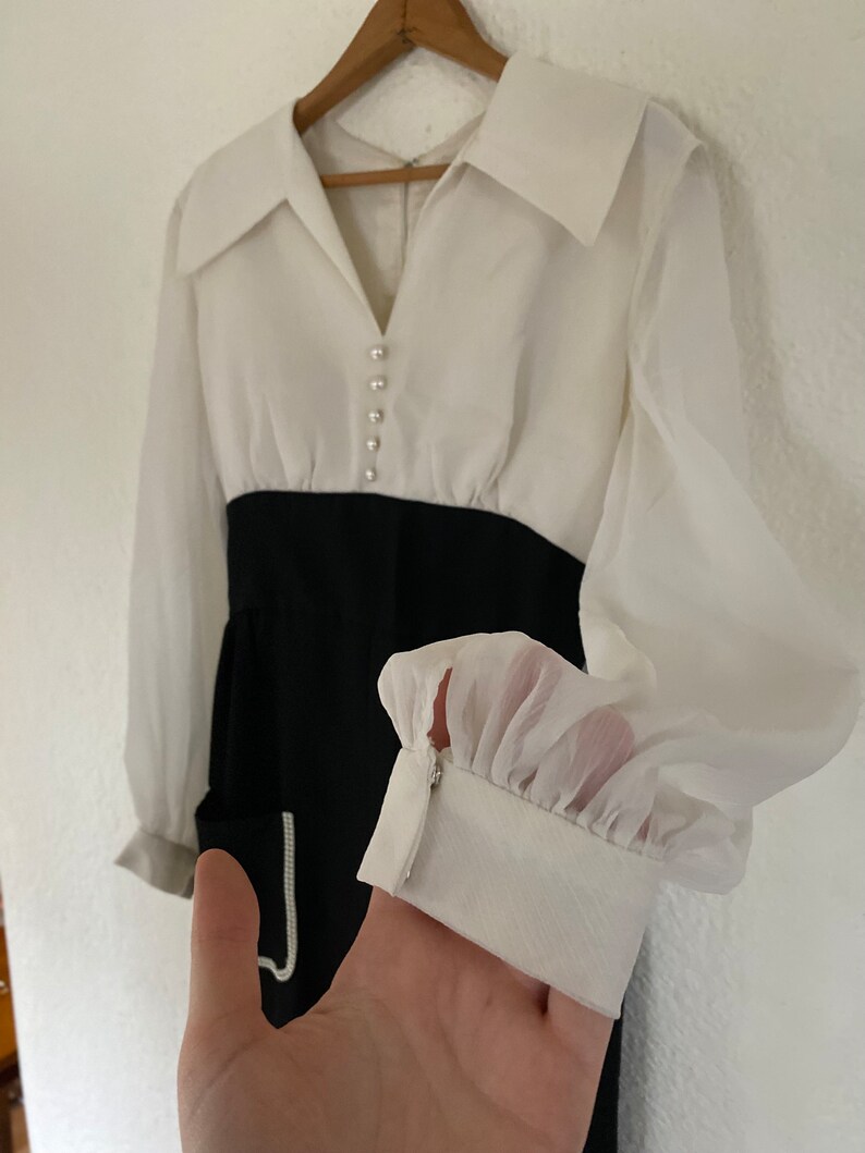 May include: A white and black dress with a large collar and long sleeves. The dress has a white top with pearl buttons and a black skirt. The sleeves are sheer and have a ruffle at the cuff.