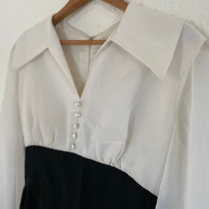 May include: A white long-sleeved blouse with a black skirt. The blouse has a large collar and a row of pearl-like buttons down the front.