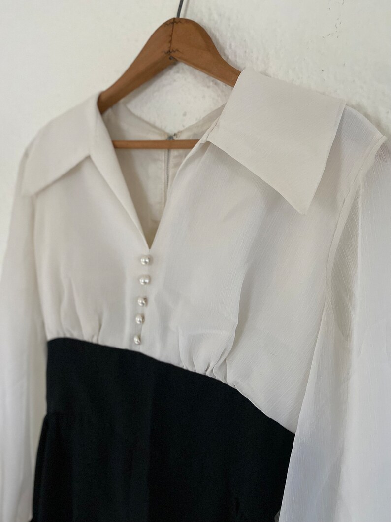 May include: A white long-sleeved blouse with a black skirt. The blouse has a large collar and a row of pearl-like buttons down the front.