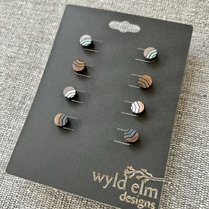May include: A set of six wooden stud earrings on a black display card. The earrings feature geometric designs in various colors, including brown, white, gray, and mint green. The card has the "wyld elm designs" logo in gold.