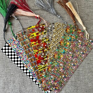 May include: A set of eight colorful bookmarks with different patterns, including checkered, confetti, books, coffee cups, bees, and camping. Each bookmark has a tassel attached.