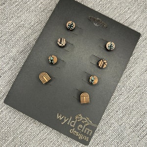 May include: A set of wooden stud earrings on a black display card. The earrings feature various designs, including leaves, geometric patterns, and rainbows. The card is labeled with the text "wyld elm designs".