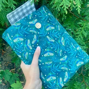 May include: A teal blue fabric pouch with a white button and a white and gray checkered fabric lining. The pouch has a pattern of white moths and green leaves on a teal blue background.