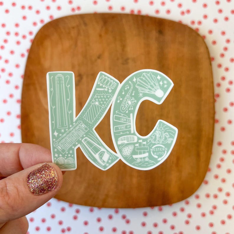 May include: A green sticker with a white outline of the letters "KC" filled with illustrations of landmarks and businesses in Kansas City, Missouri. The sticker is on a wooden surface.