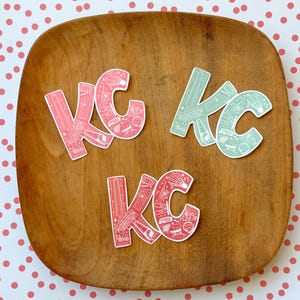 May include: Four stickers with the letters "KC" in a stylized font. The stickers are on a wooden surface. The letters are filled with a pattern of small white illustrations on a pink or green background.