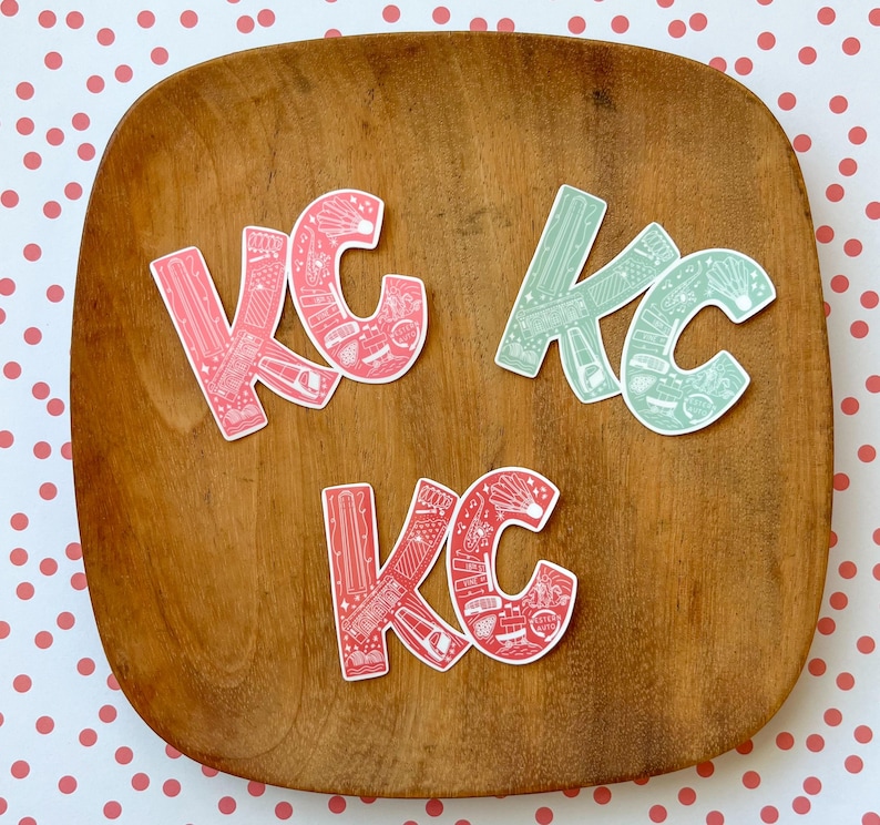 May include: Four stickers with the letters "KC" in a stylized font. The stickers are on a wooden surface. The letters are filled with a pattern of small white illustrations on a pink or green background.