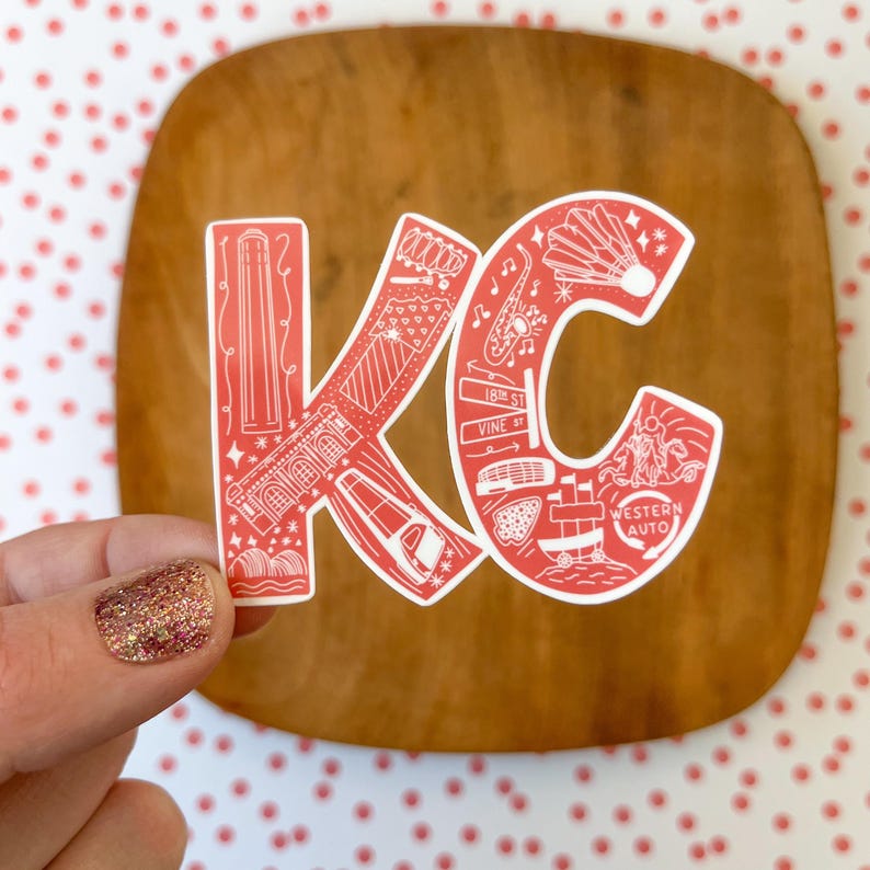 May include: Red and white sticker with the letters "KC" in a bold font. The letters are filled with illustrations of landmarks and other details from Kansas City, Missouri. The sticker is on a wooden surface.