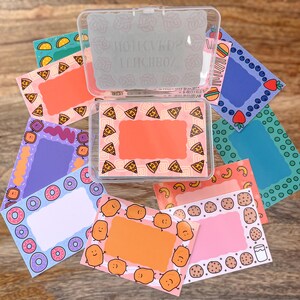 May include: A clear plastic box filled with colorful cards featuring different food themes. Each card has a white border and a blank space for writing. The themes include pizza, donuts, french fries, cookies, and blueberries.