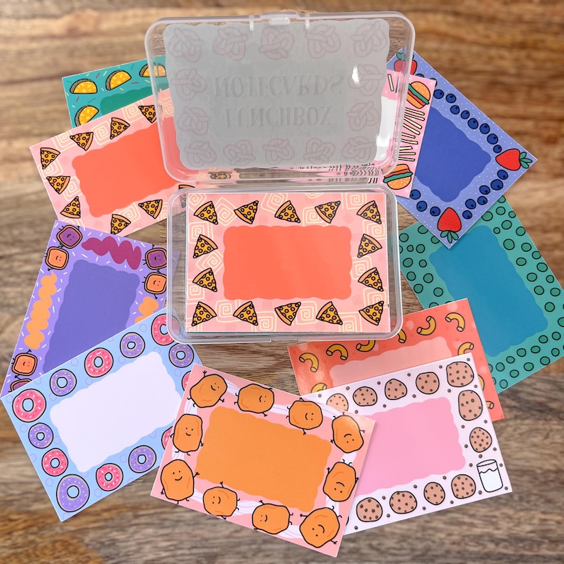 May include: A clear plastic box filled with colorful cards featuring different food themes. Each card has a white border and a blank space for writing. The themes include pizza, donuts, french fries, cookies, and blueberries.