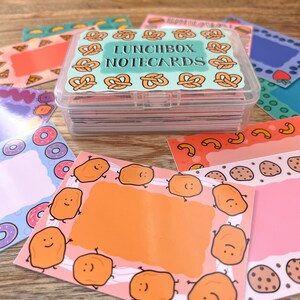 May include: A clear plastic box filled with colorful notecards. The box has a blue lid with the text "Lunchbox Notecards" printed on it. The notecards feature various designs, including pretzels, donuts, and chicken nuggets.
