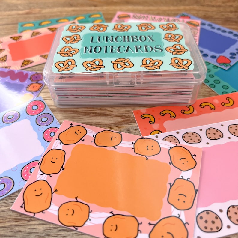 May include: A clear plastic box filled with colorful notecards. The box has a blue lid with the text "Lunchbox Notecards" printed on it. The notecards feature various designs, including pretzels, donuts, and chicken nuggets.