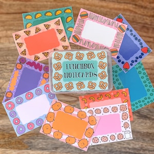 May include: A set of colorful notecards with various food-themed borders. The notecards are in a clear plastic box with the text "Lunchbox Notecards" printed on the front.