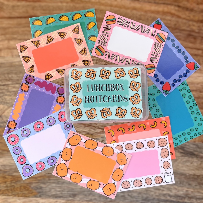 May include: A set of colorful notecards with various food-themed borders. The notecards are in a clear plastic box with the text "Lunchbox Notecards" printed on the front.