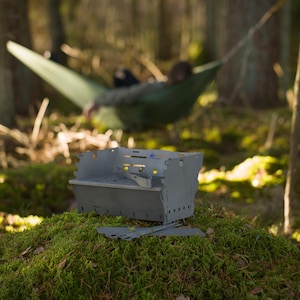 Puede incluir: Una parrilla gris, portátil y plegable, se encuentra sobre musgo verde en un entorno forestal. La parrilla es de metal cortado y tiene forma rectangular con varias aberturas. Una pieza separada se encuentra cerca. Una persona se relaja en una hamaca en el fondo.