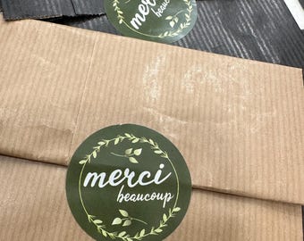 May include: Two packages, one black and one brown, are sealed with round green stickers. The stickers have the words "merci beaucoup" in white script, surrounded by a wreath of leaves. The packages are on a black and white checkered surface.