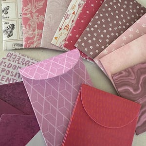 May include: A selection of decorative envelopes in shades of pink and red. The envelopes display a variety of patterns, including floral, geometric, and arrow designs. Some envelopes feature text and butterfly illustrations.