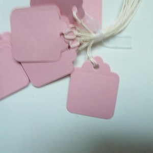 Può includere: Un mucchio di etichette regalo di carta rosa con uno spago bianco attaccato. Le etichette hanno un bordo a festoni e sono vuote.