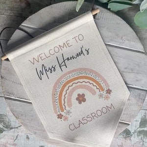 K&ouml;nnte beinhalten: Ein beigefarbenes Canvas-Banner mit dem Text "Willkommen in Miss Harwoods Klassenzimmer" und einem Regenbogen mit Blumen in Rosa-, Orange- und Braunt&ouml;nen.