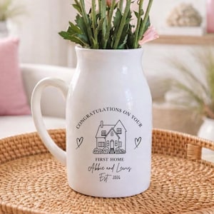 May include: A white ceramic pitcher vase filled with pink chrysanthemums. The front features black text reading CONGRATULATIONS ON YOUR FIRST HOME, Abbie and Lewis, EST 2026, a line drawing of a house, and two small heart icons on either side.