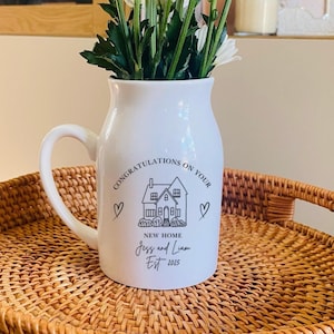 May include: A white ceramic pitcher filled with white daisy flowers. The pitcher features black text that reads CONGRATULATIONS ON YOUR NEW HOME, Jess and Liam, Est 2025, along with a line drawing of a house and two small heart symbols on the front.