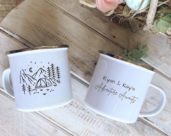 May include: Two white enamel camping mugs with gold rims. One mug features a black illustration of a tent, mountains, and trees. The other mug has the text "Ryan & Kay's Adventure Awaits" in black script. The mugs are on a wooden surface.