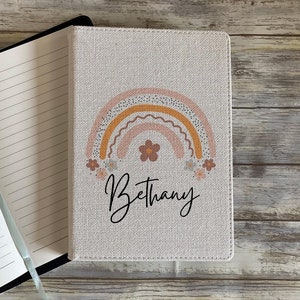 May include: A beige linen journal with a rainbow design and the name "Bethany" printed on the front cover.
