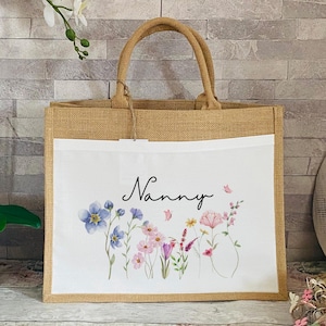 K&ouml;nnte beinhalten: Eine nat&uuml;rliche Jute-Tragetasche mit einem wei&szlig;en Stoffpanel, das mit einem Blumendesign und dem Wort "Nanny" in schwarzer Schrift verziert ist.