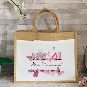 May include: A natural jute tote bag with a white canvas insert featuring a pink and black design of school supplies, butterflies, and the text "Mrs. Stevens".