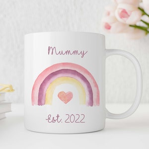 May include: White ceramic mug with a pink and purple watercolor rainbow design. The rainbow has a small pink heart in the center. The text "Mummy est. 2022" is printed below the rainbow.