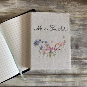 May include: A notebook with the name "Mrs Smith" in script on the cover. The cover has a floral design with blue, pink, and purple flowers. The notebook is open, showing lined pages and a blue ribbon.