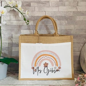 May include: A brown burlap tote bag with a white insert featuring a rainbow design with pink, orange, and yellow colors. The text "Mrs Gibson" is printed below the rainbow.