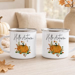 May include: Two white enamel mugs with a silver rim, each featuring a graphic of an orange pumpkin surrounded by autumn leaves and the black cursive text Hello Autumn. The mugs have a side handle and a smooth, glossy finish on the exterior surface.