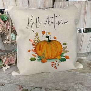 May include: A square off-white throw pillow featuring the black cursive text Hello Autumn above a central illustration of a bright orange pumpkin surrounded by colorful fall foliage, including green, yellow, and orange leaves and small berries.