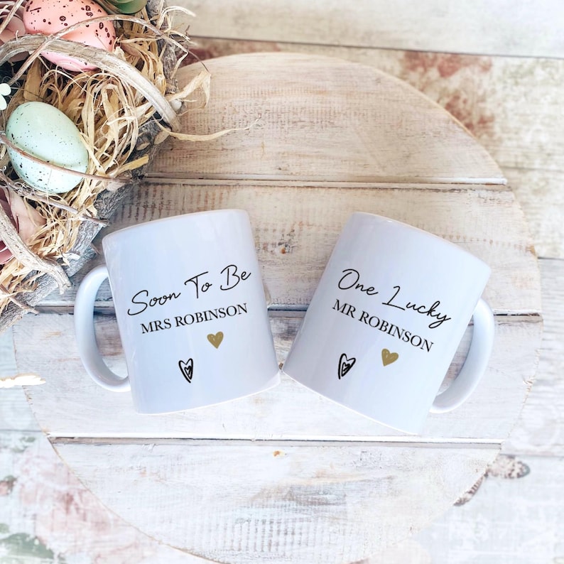 May include: Two white ceramic mugs with black text and gold heart details. One mug says "Soon To Be MRS ROBINSON" and the other says "One Lucky MR ROBINSON". Each mug features a black heart design.