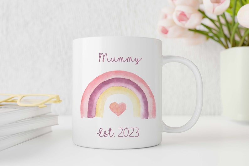 May include: White ceramic mug with a pink and purple watercolor rainbow design. The rainbow has a small pink heart in the center. The text "Mummy est. 2023" is printed below the rainbow.